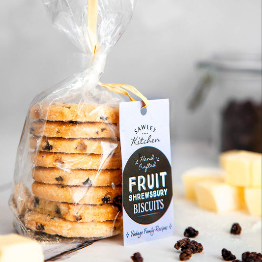 Packaged Fruit Shrewsbury Biscuits in a clear plastic bag with a label showing the brand and product name, along with some loose biscuits and dried currants on the table.