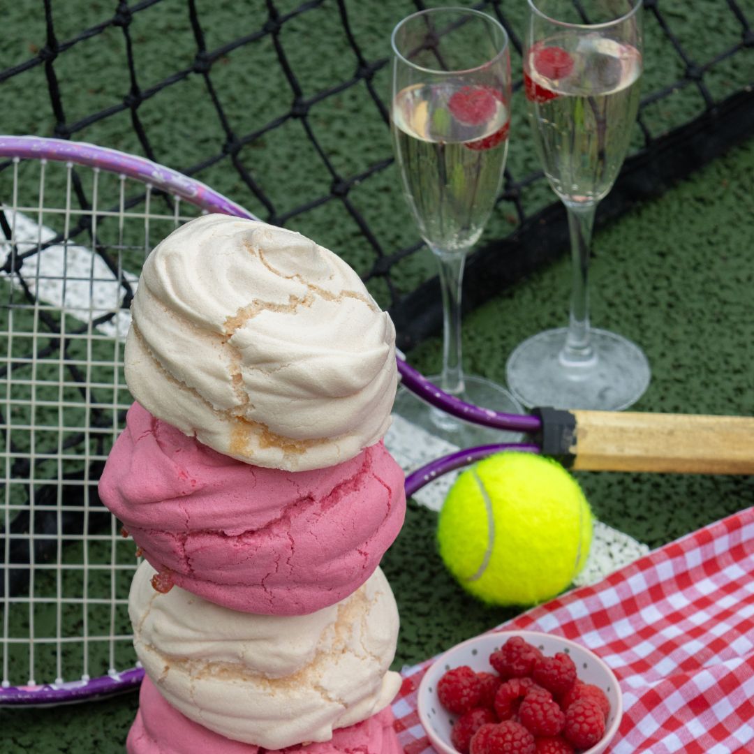 Stack of vanilla and raspberry meringues with a tennis racket, ball, and glasses on a tennis court.