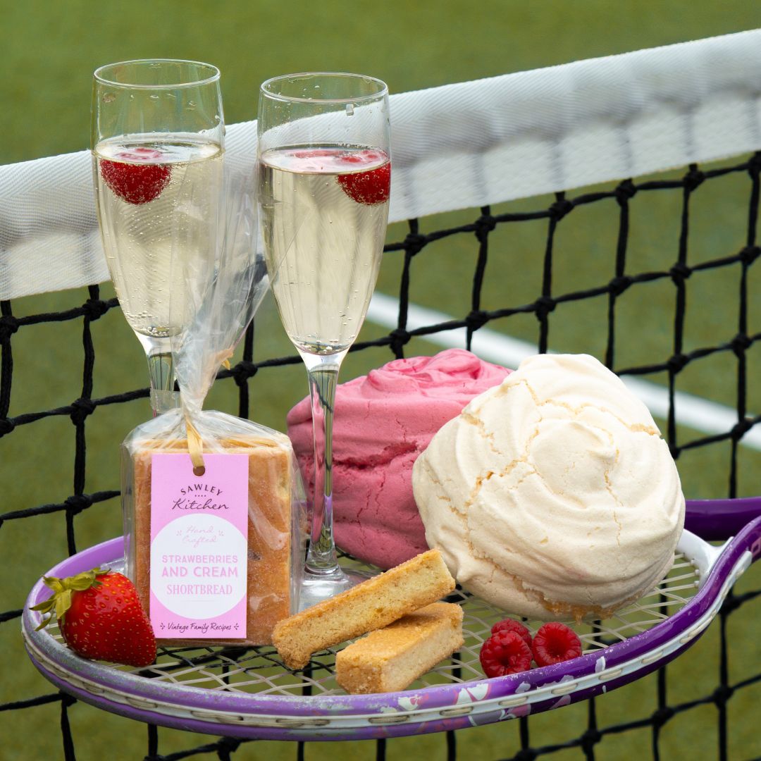 Two champagne glasses with raspberries, a bag of strawberries and cream shortbread, a vanilla meringue and a raspberry meringue and tennis racket against a tennis court background.