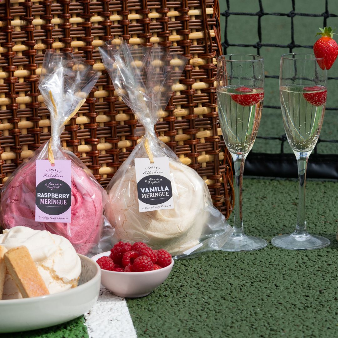 Two wrapped desserts labeled 'Raspberry Meringue' and 'Vanilla Meringue' with two glasses of champagne on a tennis court-themed setting.
